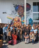 a large, diverse group of smiling people posing for a photo in front of a colorful mural of a woman's face