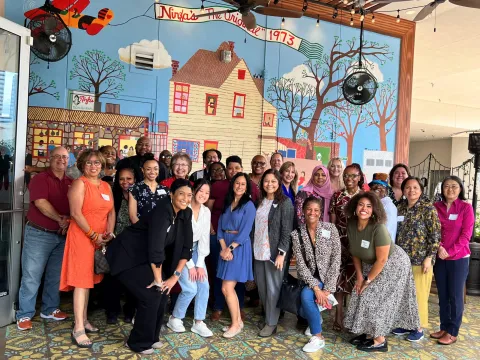 A group of 27 people pose for a photo in front of a mural marked "Ninfa's The Original 1973," suggesting a gathering at a historical or significant location. The diverse group, displaying a mix of ages, races, and attire, seems to be at a social or professional event, indicated by their casual to business-casual clothes and name tags. They stand in a well-lit indoor space that appears to be a restaurant or public venue.