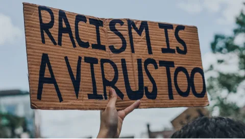 Hand holding up a cardboard sign that says, "Racism is a Virus Too"