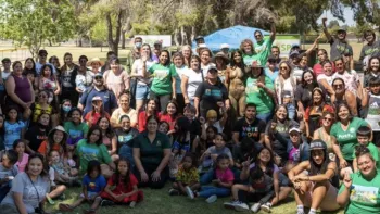 The image shows a large group of people gathered outdoors for what appears to be a community event or social gathering in a park setting. The group is diverse in terms of age and gender, with both children and adults present, many of whom are smiling and posing for the photo. Some individuals are standing while others are seated on the grass. 