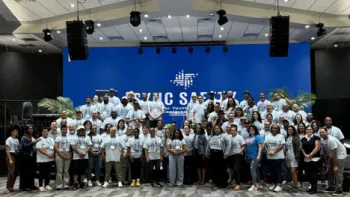 A large group of diverse individuals standing on a stage and posing for a group photo. They are all wearing matching light blue t-shirts, and the stage backdrop features a blue wall with the text "SYNC SOUTH: Better Together Youth & Family Engagement." The setting appears to be a spacious, modern hall with bright overhead lighting and large screens on either side of the stage.